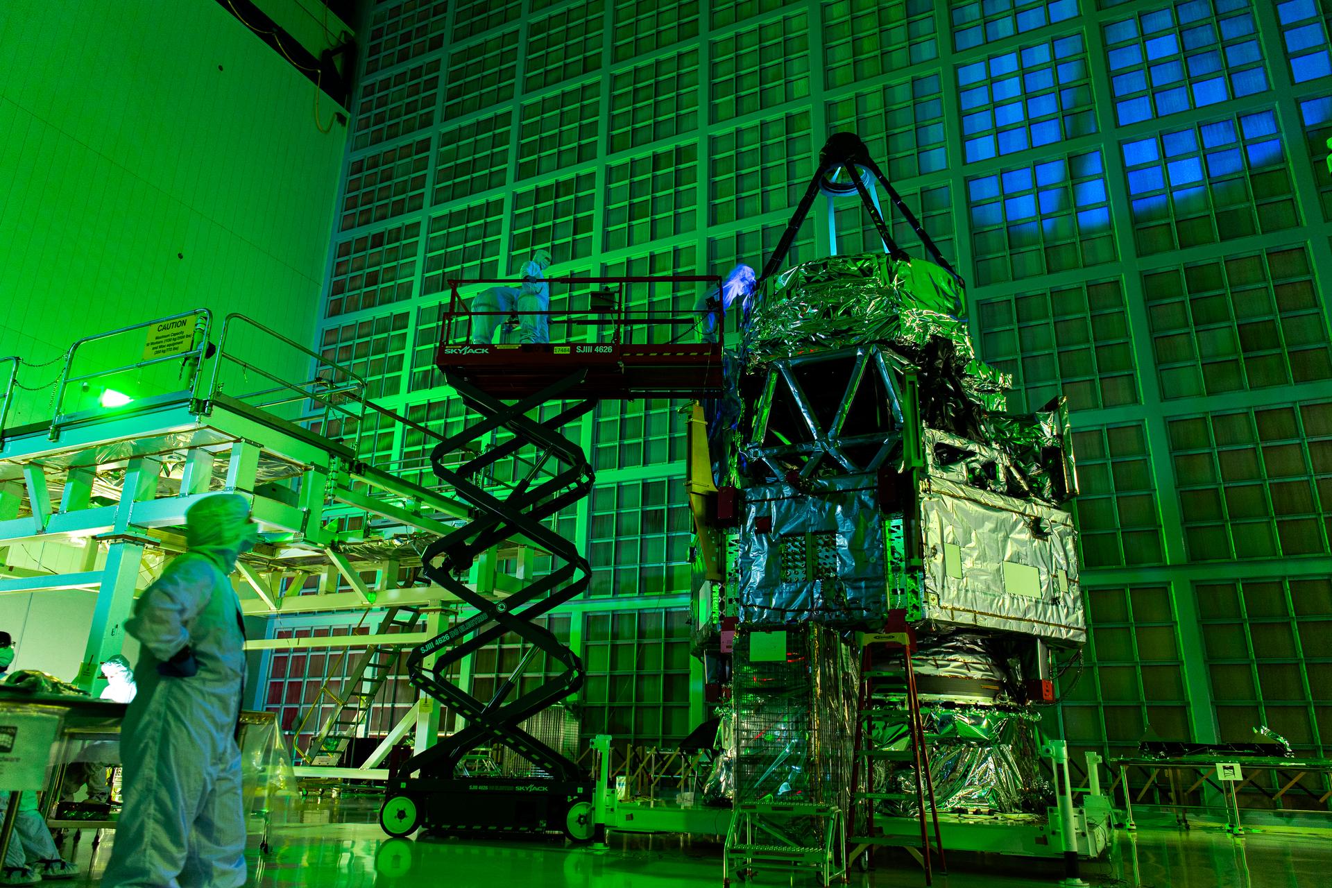 Engineers in cleanroom suits inspect the Roman Space Telescope under green laser light at NASA Goddard Space Flight Center